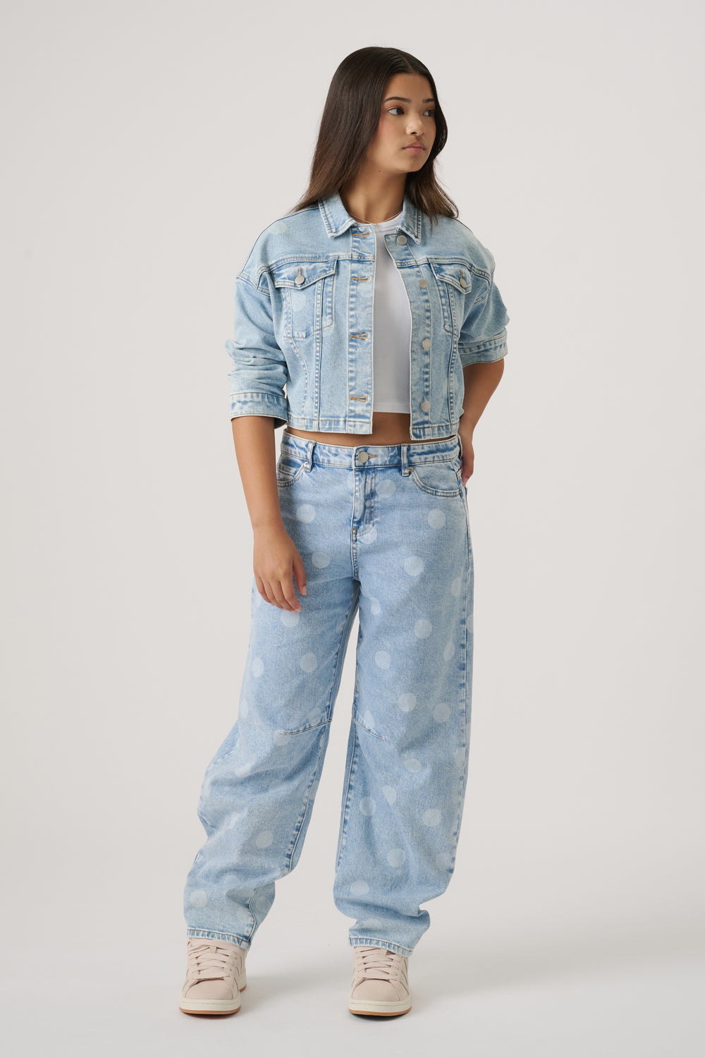 Teen girl wearing a light blue cropped denim jacket over a white crop top with wide-leg polka dot jeans and sneakers