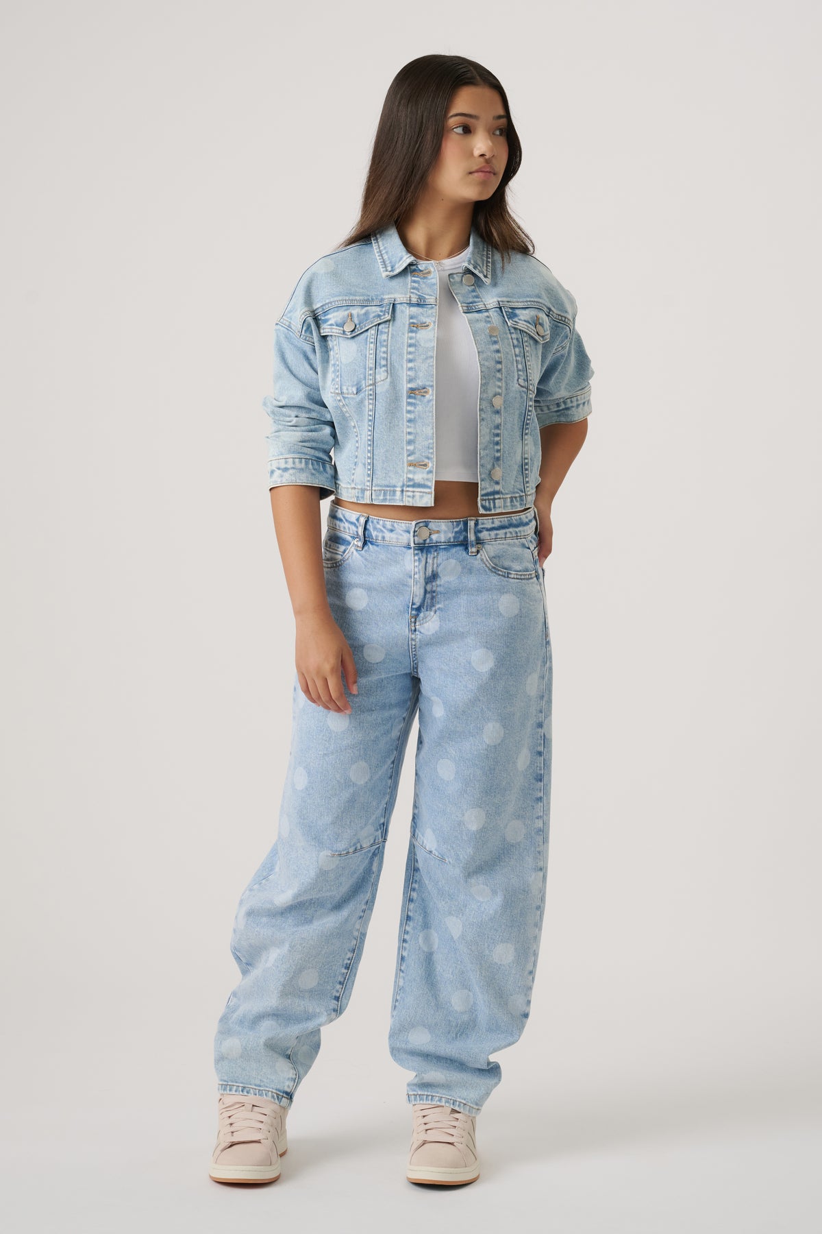 Teen girl wearing a light blue cropped denim jacket over a white crop top with wide-leg polka dot jeans and sneakers