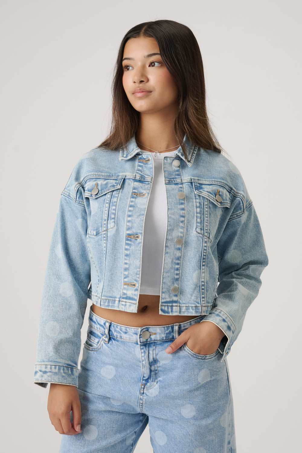 Teen girl wearing a light wash cropped denim jacket with wide sleeves over a white crop top and matching jeans