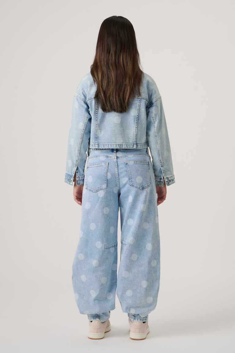 Girl wearing a cropped light-wash denim jacket paired with wide-leg polka dot jeans and cream sneakers, viewed from behind