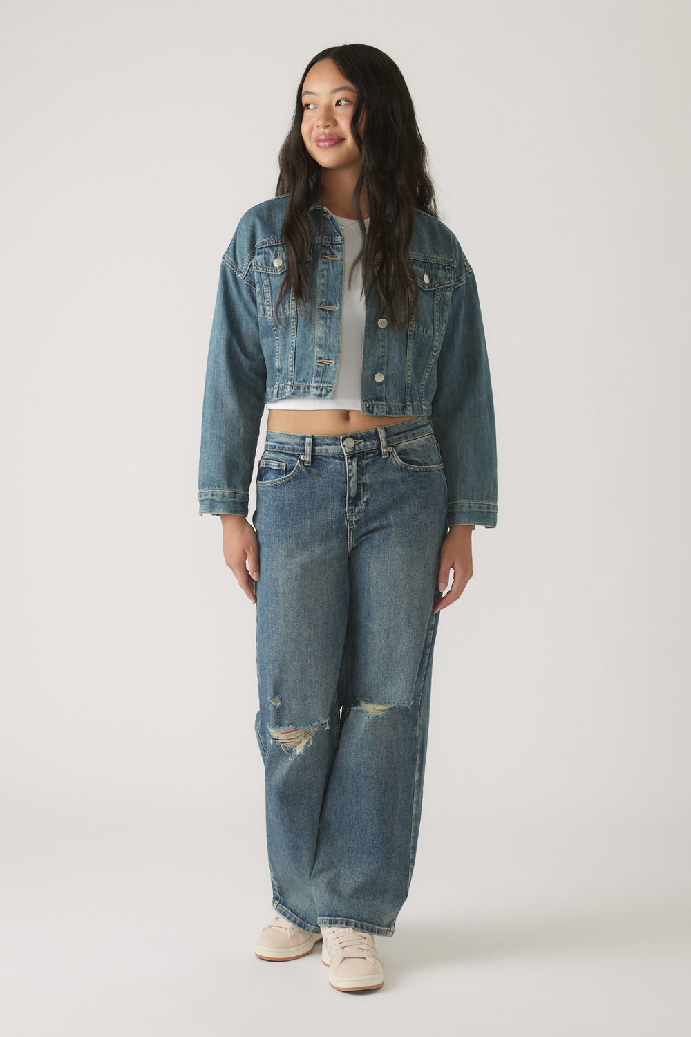 Teen girl wearing a cropped denim jacket layered over a white top with relaxed ripped jeans and casual sneakers