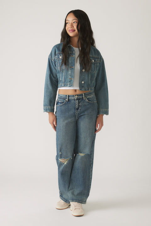 Teen girl wearing a cropped denim jacket layered over a white top with relaxed ripped jeans and casual sneakers