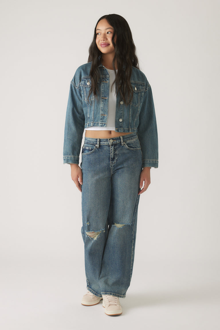 Teen girl wearing a cropped denim jacket layered over a white top with relaxed ripped jeans and casual sneakers