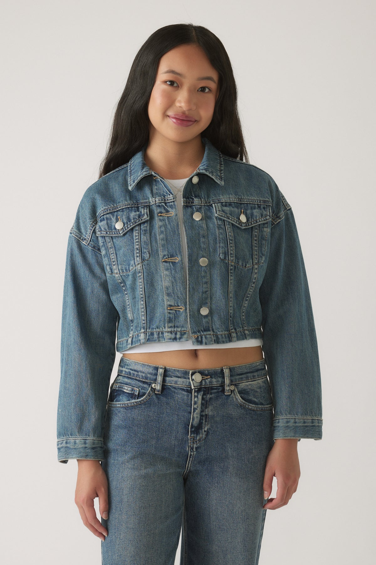 Girl wearing a mid-wash cropped denim jacket with front pockets, layered over a white cropped top and denim jeans