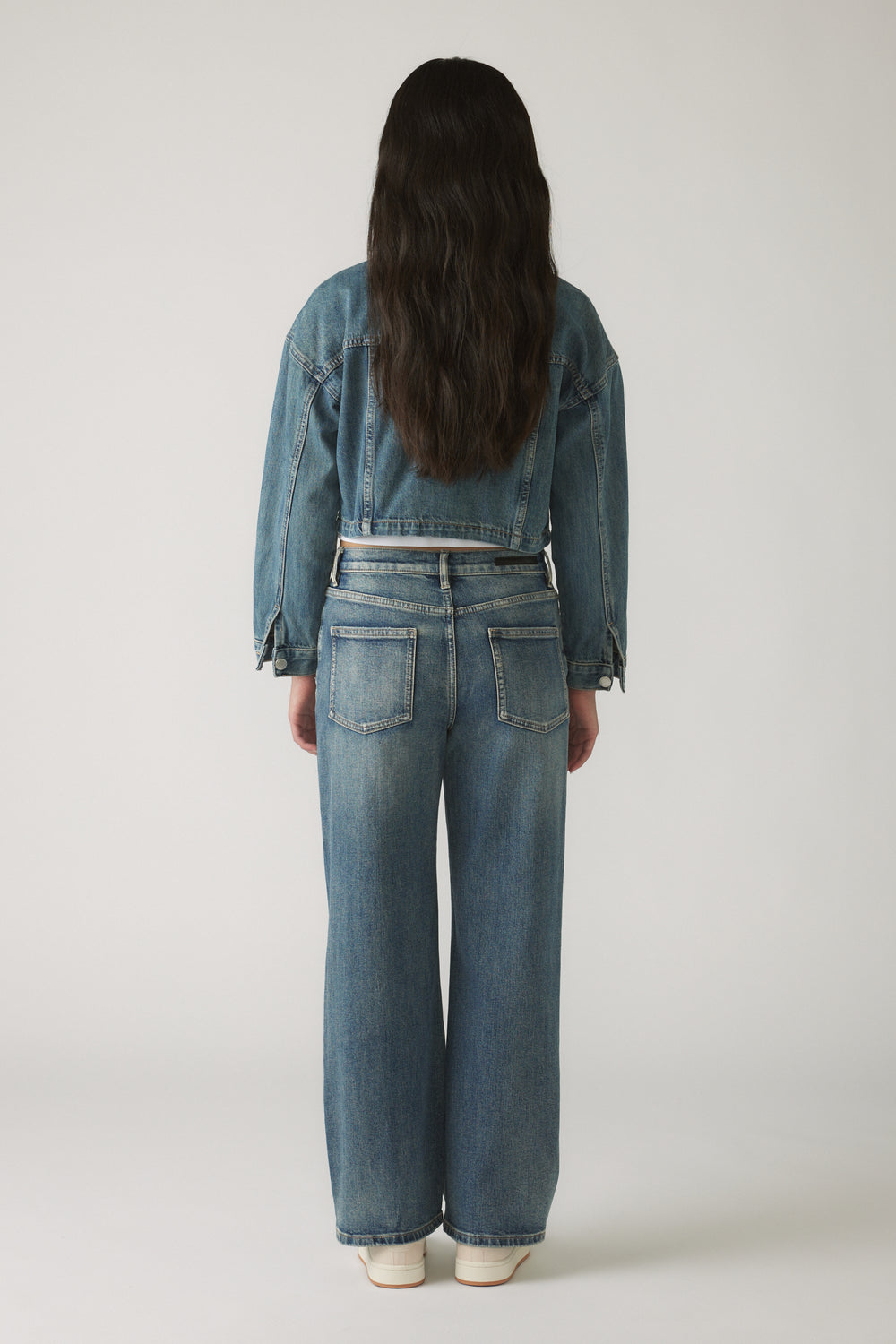 Back view of a girl wearing a cropped blue denim jacket paired with straight leg denim jeans and white sneakers