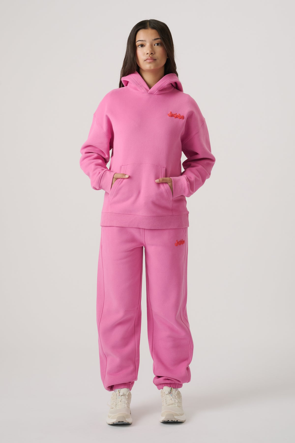 Girl wearing bright pink balloon leg trackpants paired with matching hoodie and white sneakers standing against neutral background.