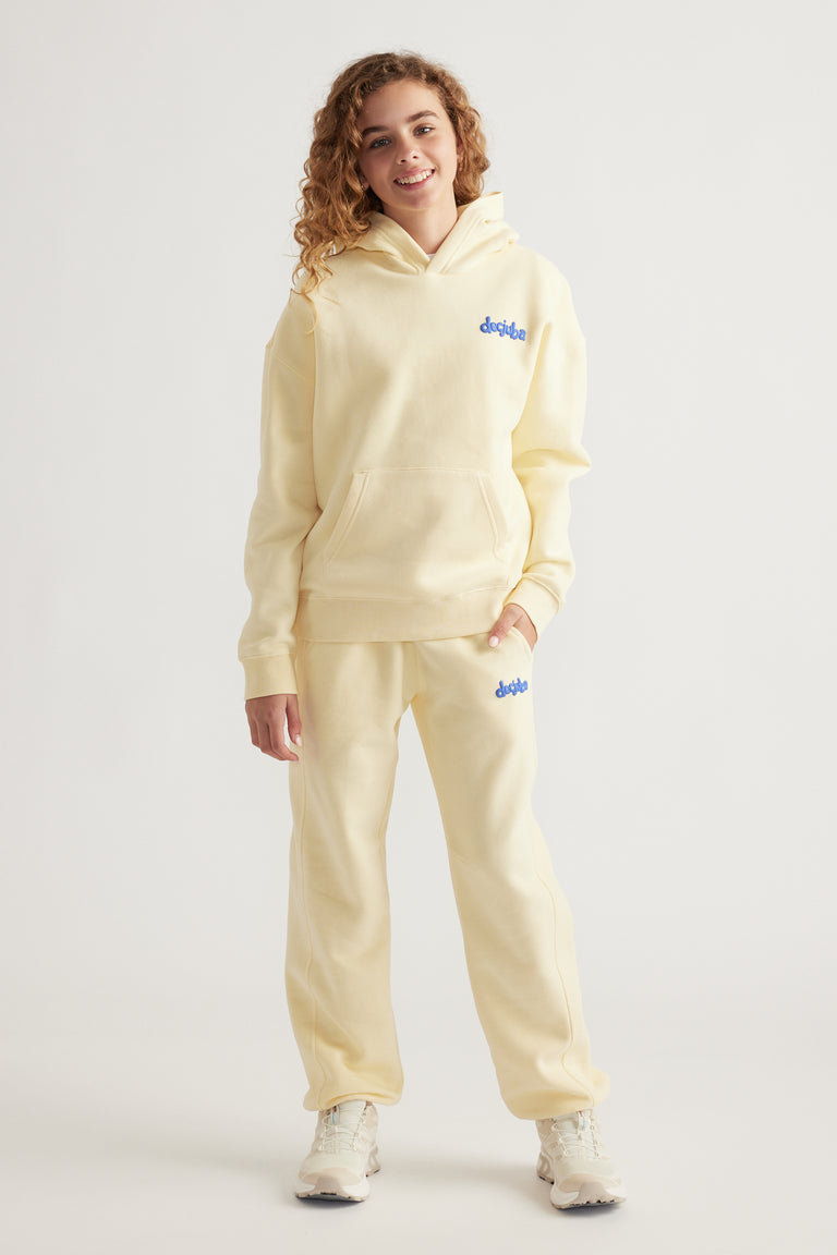 Teen girl wearing soft yellow hoodie and matching relaxed pants with subtle blue logo, styled with white sneakers