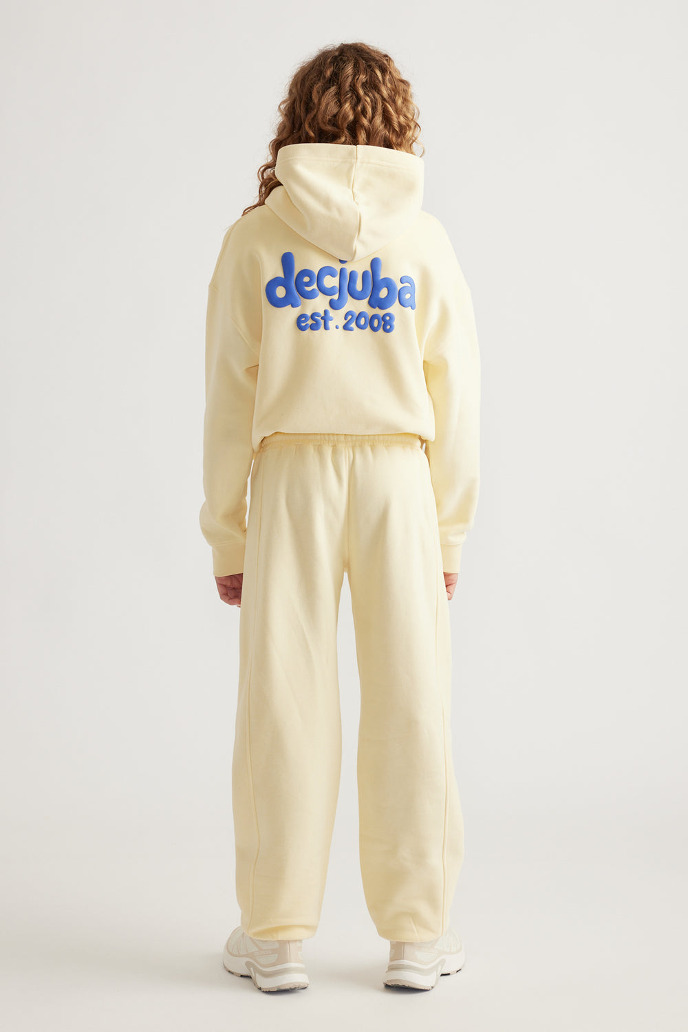 Teen girl wearing a cream hooded sweatshirt with blue logo and matching relaxed fit track pants with cuffed hems