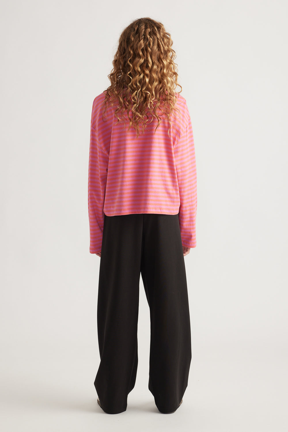 Child with curly hair wearing a relaxed fit pink and orange striped long sleeve tee and wide black pants standing facing away