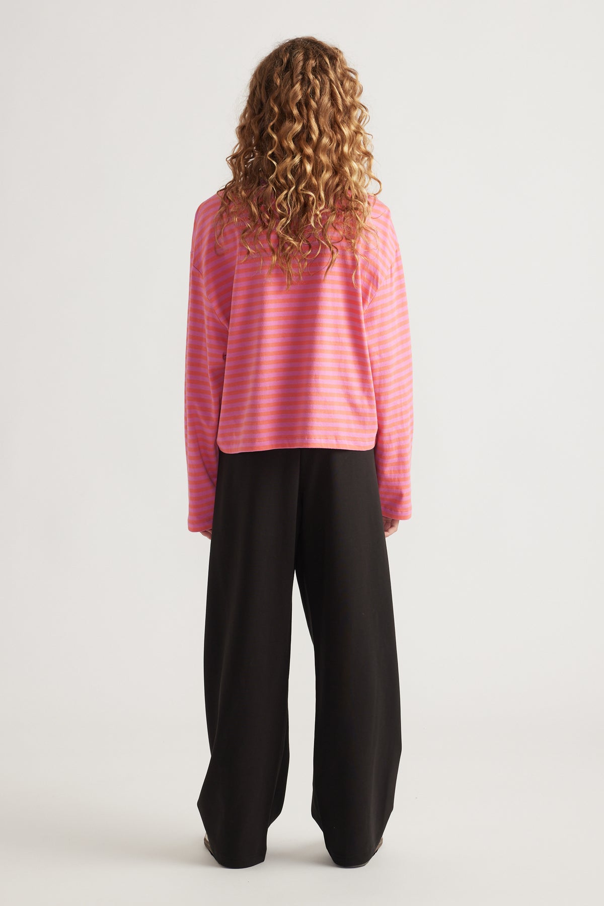Child with curly hair wearing a relaxed fit pink and orange striped long sleeve tee and wide black pants standing facing away