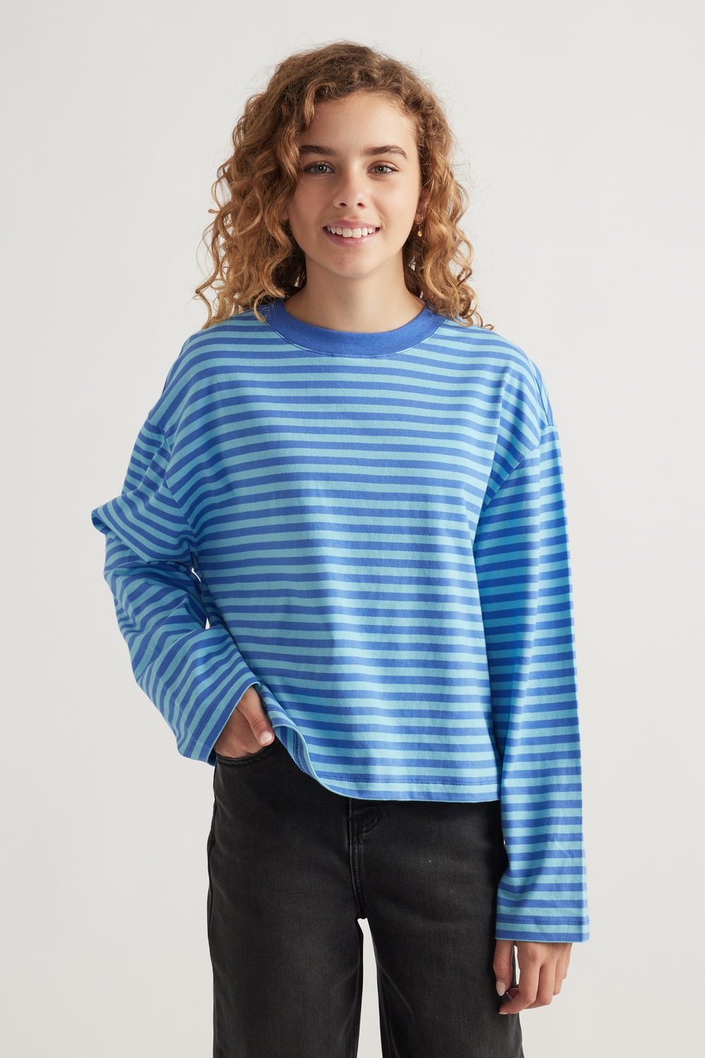 Smiling girl wearing a relaxed fit, striped blue long sleeve tee paired with black pants in a neutral studio setting
