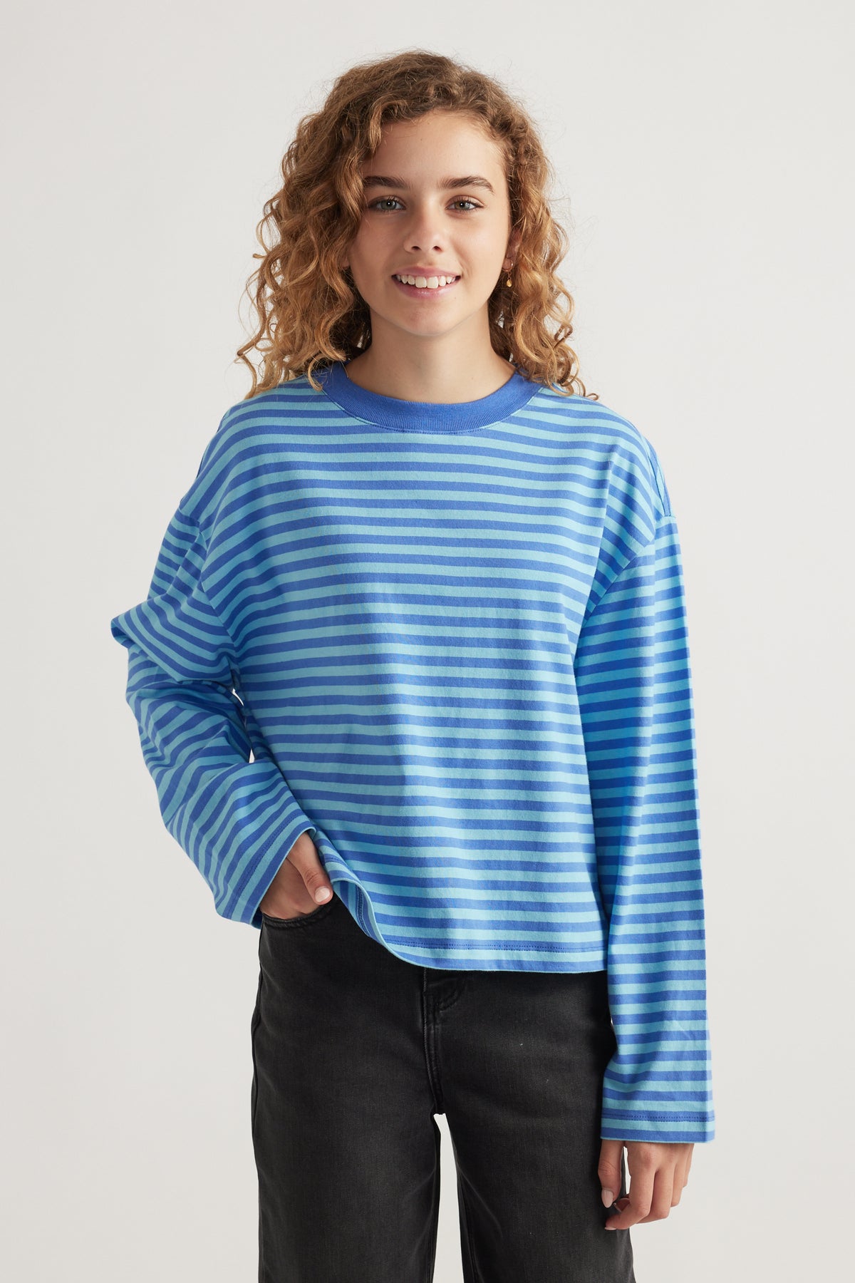Smiling girl wearing a relaxed fit, striped blue long sleeve tee paired with black pants in a neutral studio setting