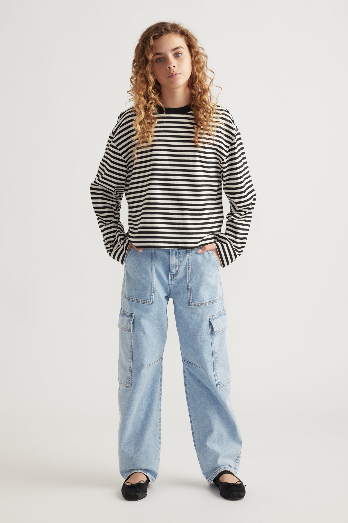 Teen girl wearing light wash barrel leg cargo jeans paired with a black and white striped long-sleeve top and flat shoes