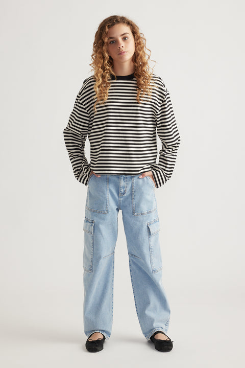 Teen girl wearing light wash barrel leg cargo jeans paired with a black and white striped long-sleeve top and flat shoes