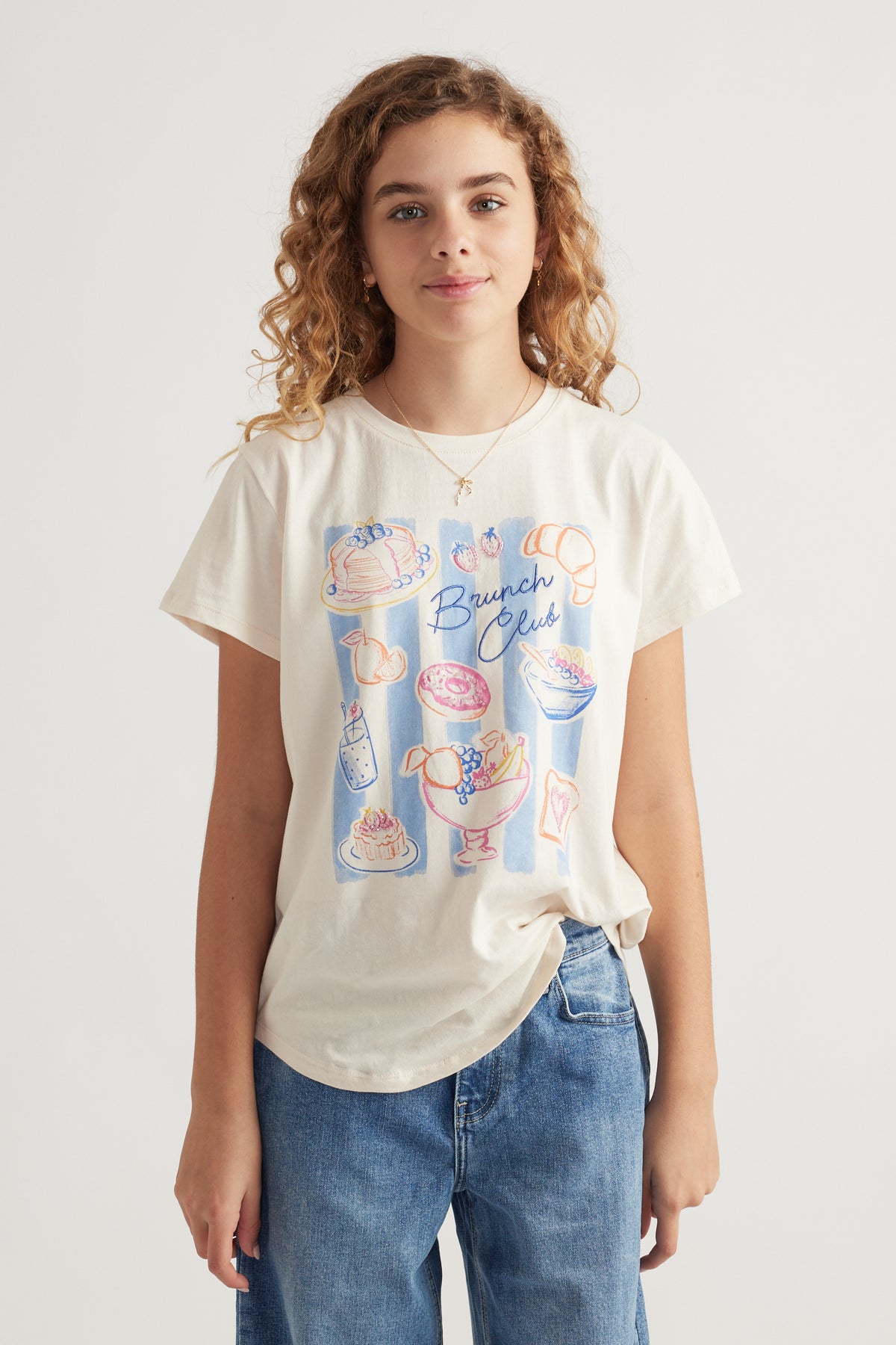 Front view of girl wearing light cream graphic tee with brunch-themed print paired with relaxed fit blue jeans