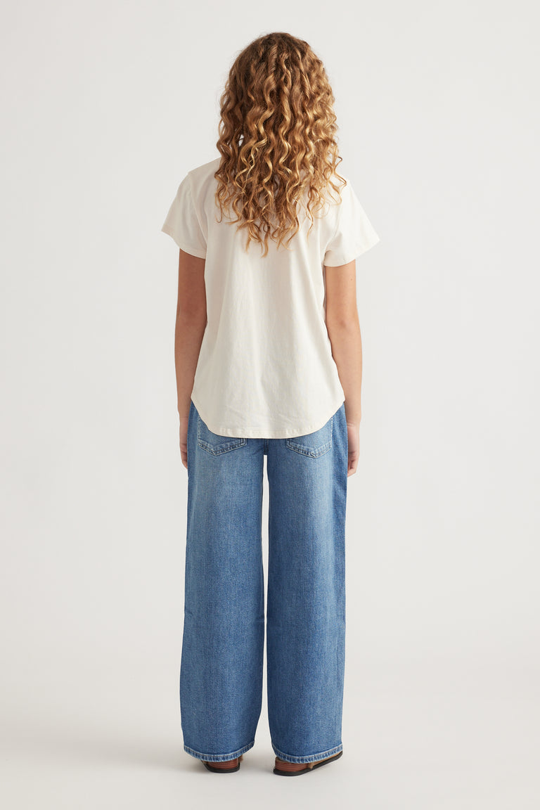 Rear view of girl wearing cream short sleeve tee with curved hem paired with wide leg light wash denim jeans