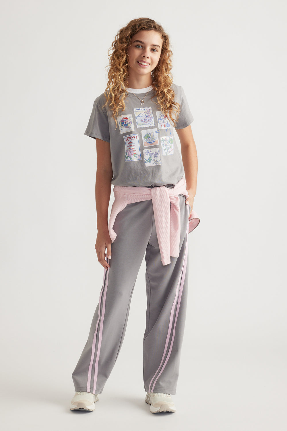 Girl wearing a printed short sleeve tee with city-themed artwork paired with grey track pants and pink sweater tied at waist