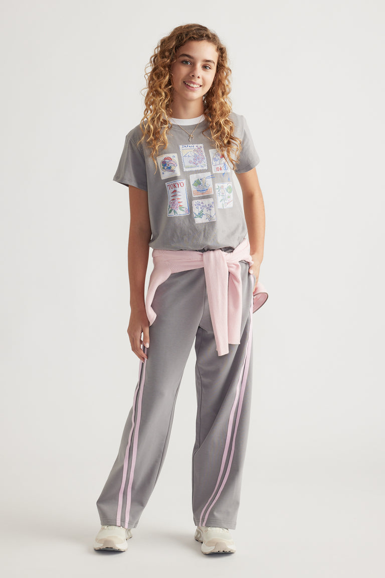 Girl wearing a printed short sleeve tee with city-themed artwork paired with grey track pants and pink sweater tied at waist