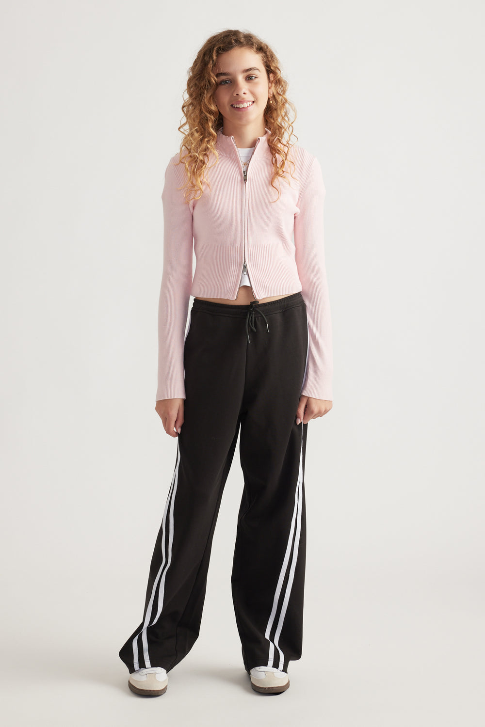 Teen girl wearing black wide-leg track pants with white side stripes and a pale pink zip-up top