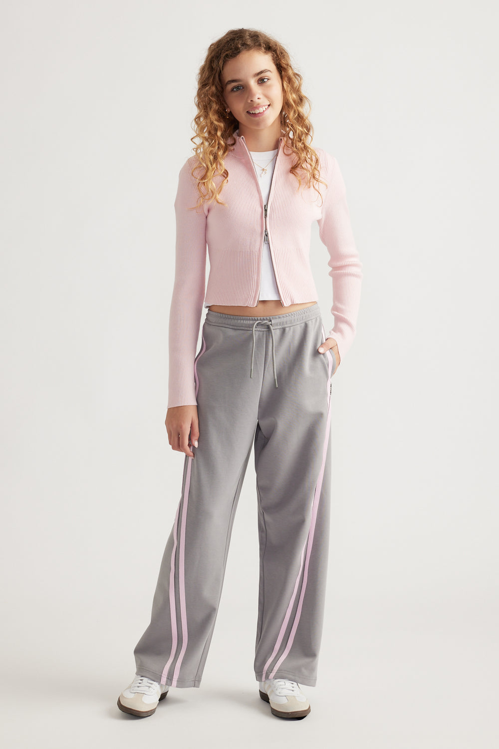 Teen girl wearing grey wide-leg pants with pink side stripes and a pale pink zip-up cardigan over white top
