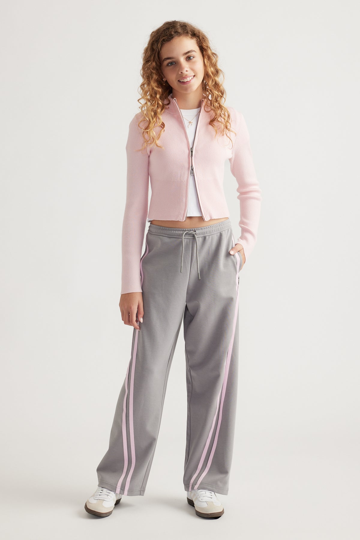 Teen girl wearing grey wide-leg pants with pink side stripes and a pale pink zip-up cardigan over white top