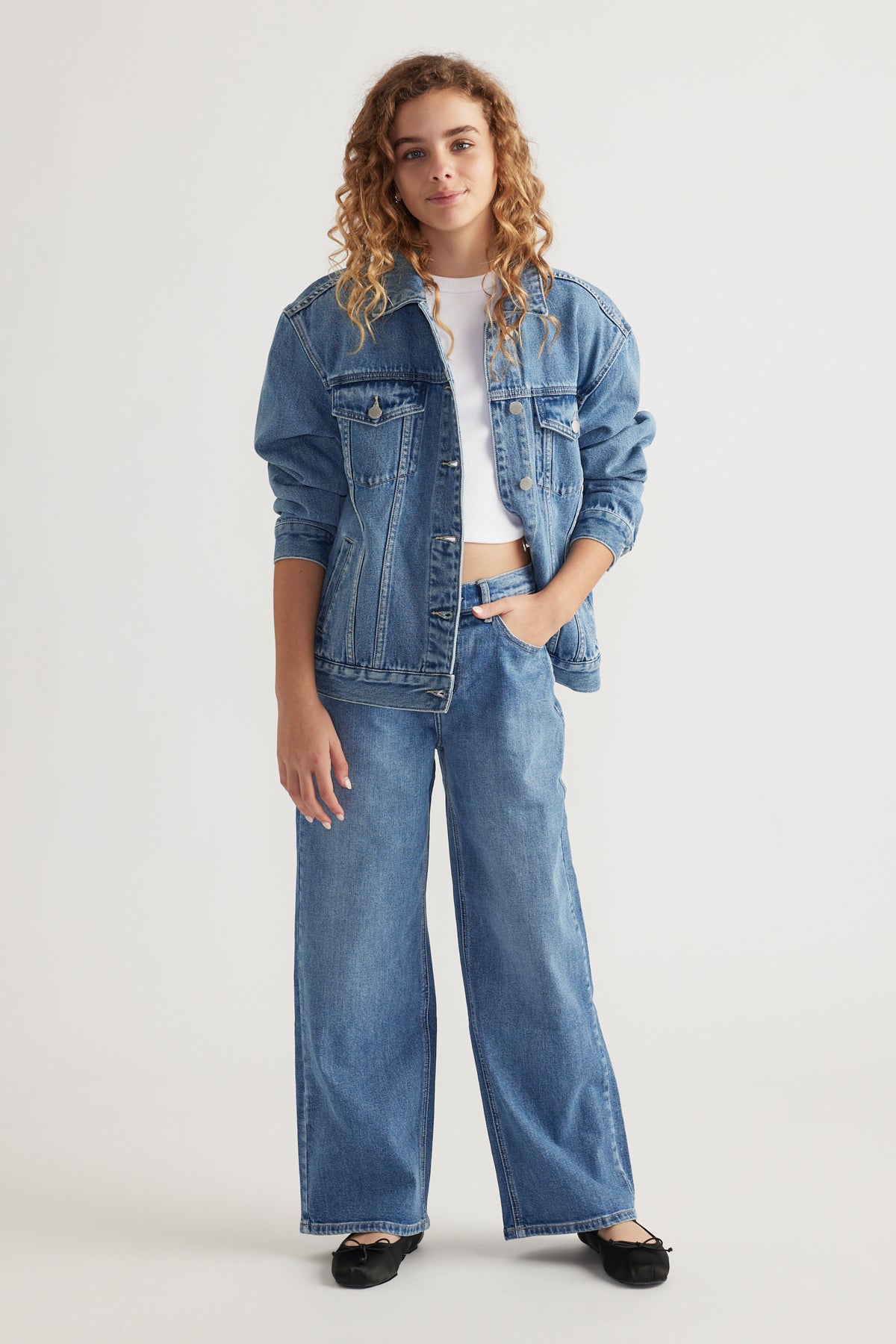Teen girl wearing oversized denim jacket layered over white crop top with wide leg denim jeans and black flats