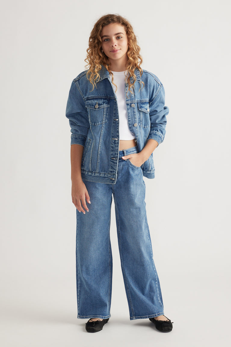 Teen girl wearing oversized denim jacket layered over white crop top with wide leg denim jeans and black flats