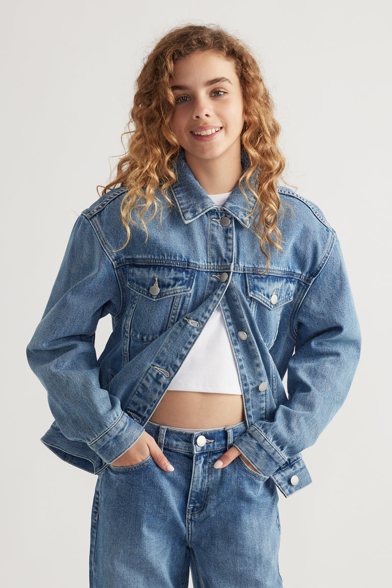 Teen girl smiling wearing oversized blue denim jacket over white cropped top and matching denim jeans