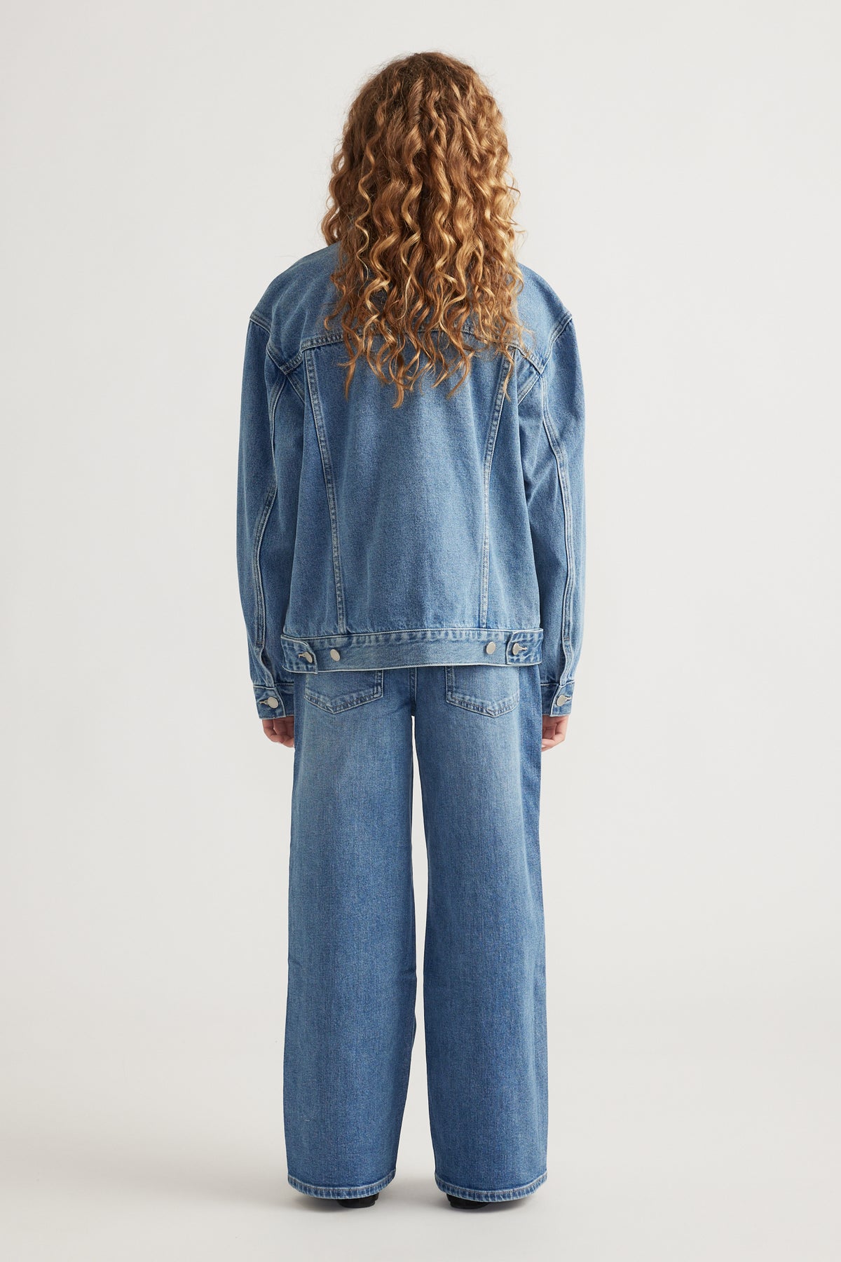 Rear view of teen girl wearing oversized denim jacket paired with wide leg denim jeans against plain background