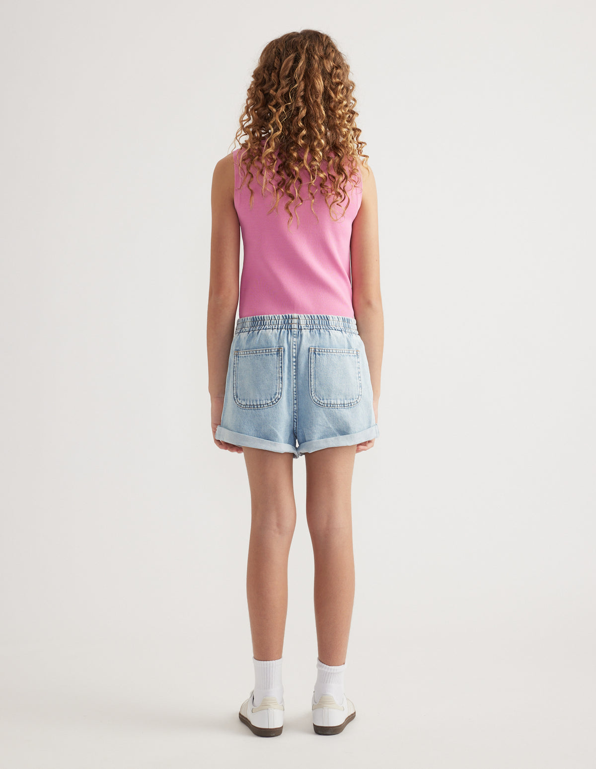 Back view of girl wearing light wash denim pull-on shorts with rolled hems, a pink tank top and sneakers