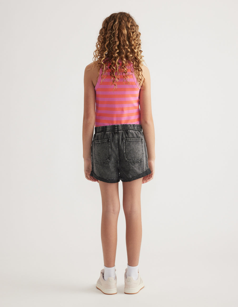 Back view of girl wearing black denim pull-on shorts with an elastic waist and cuffed hems, a striped tank and sneakers