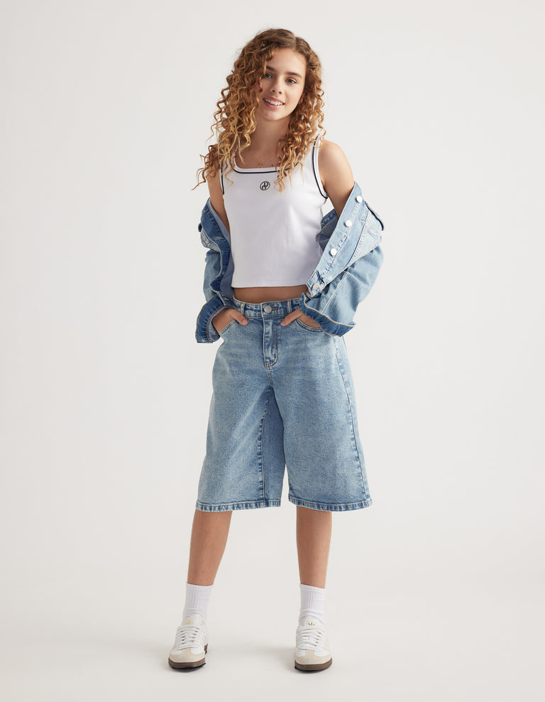 Girl wearing wide leg denim jorts with a white cropped tank top, denim jacket and sneakers