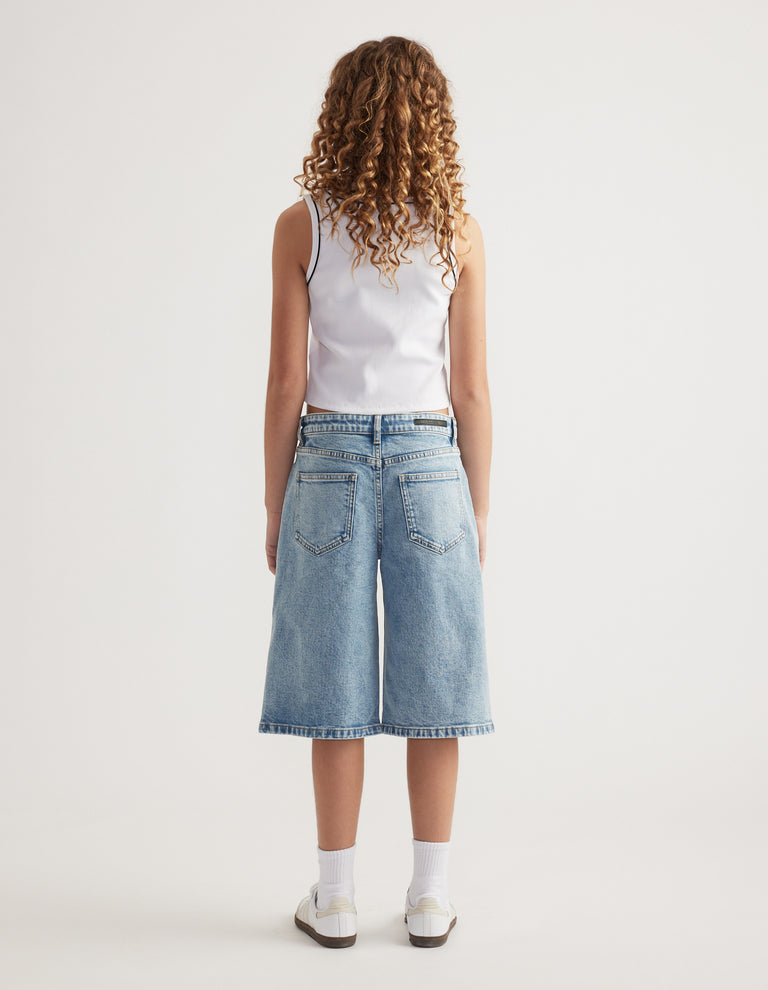 Back view of girl wearing light wash wide leg denim jorts with a white tank top and sneakers