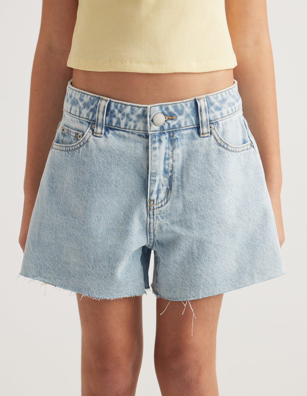 Girl wearing light wash mid rise A line denim shorts with frayed hems and a cream ribbed tank top