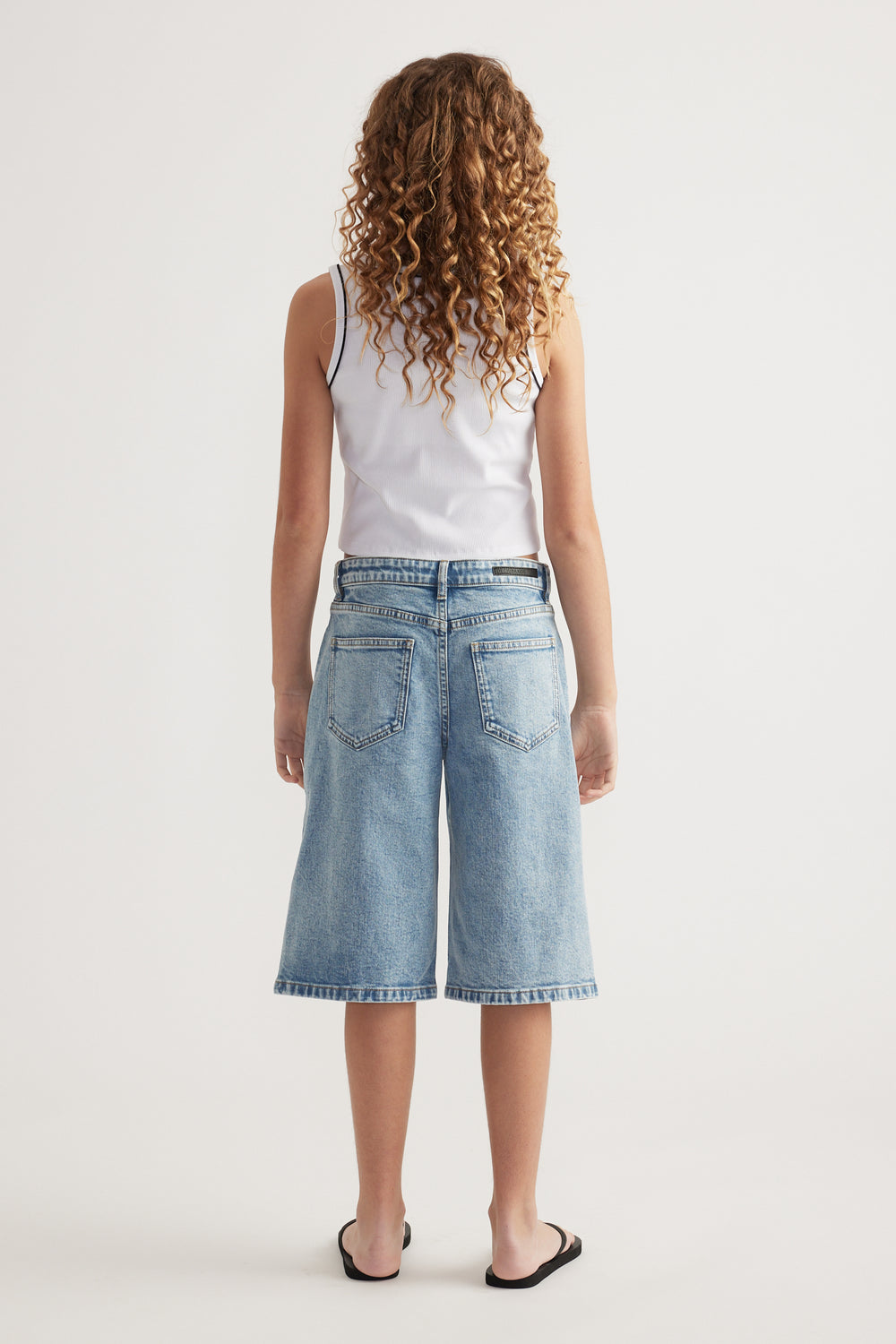 Girl seen from behind in a white ribbed scoop neck tank, wide leg denim shorts and sandals