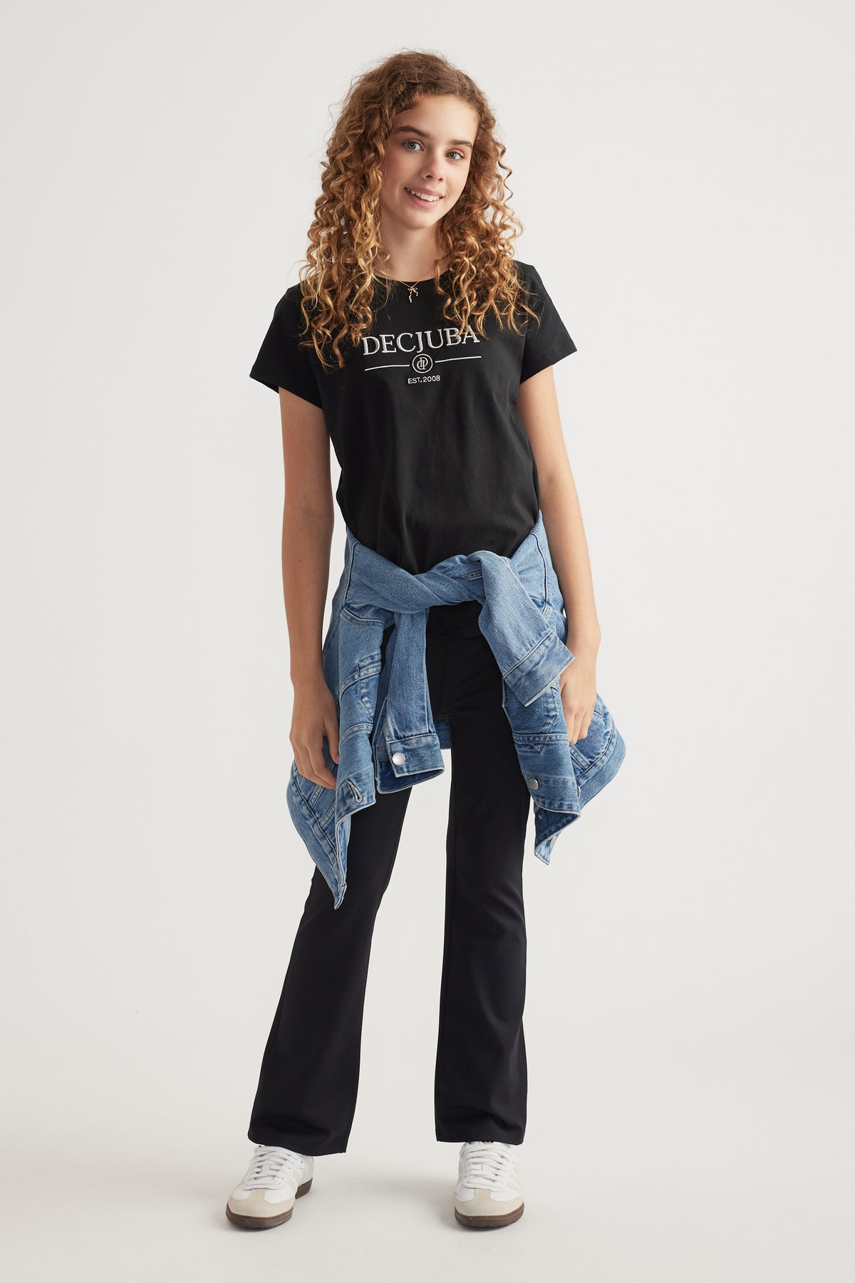 Girl wearing a black Decjuba graphic tee with black pants, a denim jacket tied at the waist and sneakers