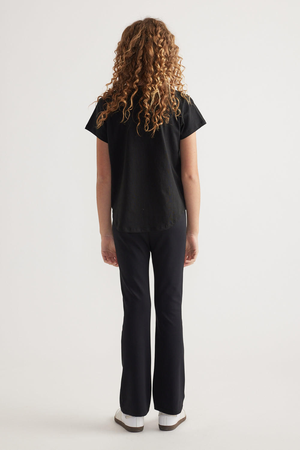 Back view of girl wearing a black heritage graphic T-shirt with black pants and sneakers