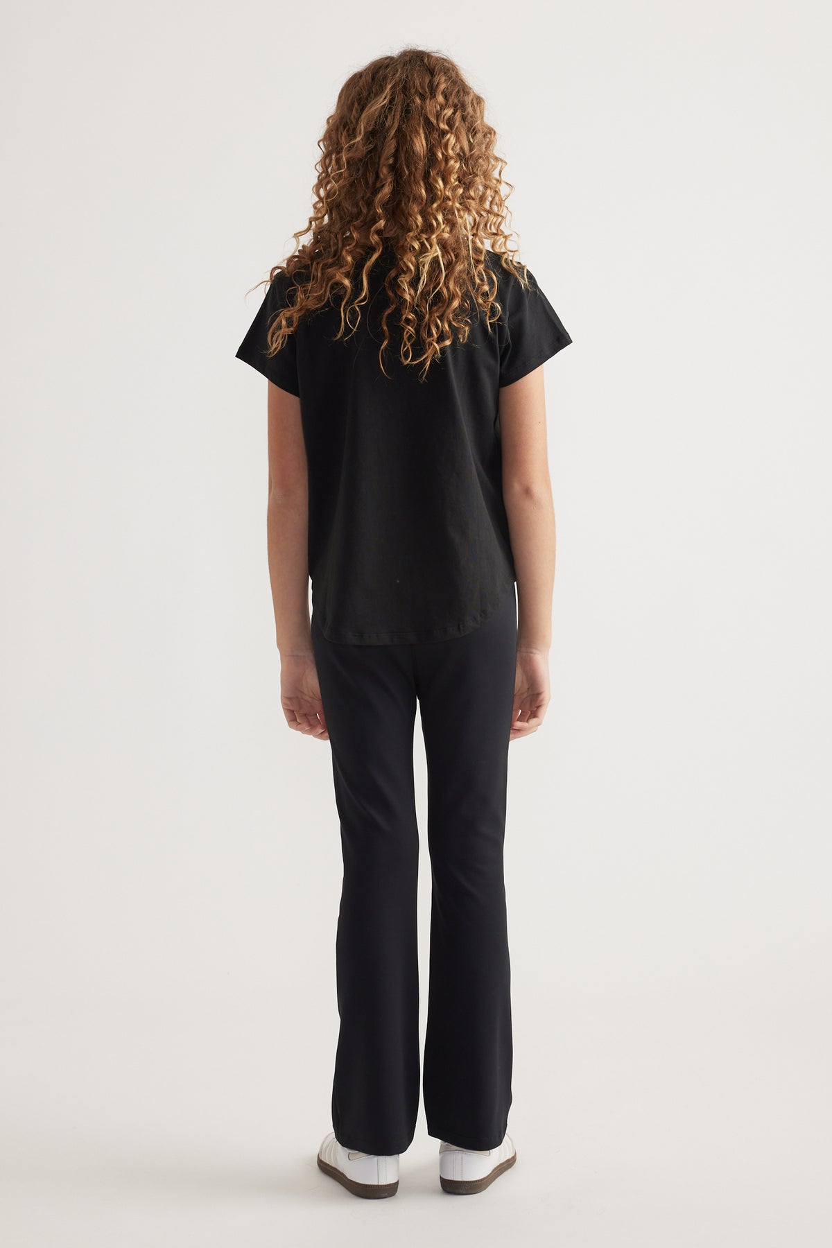 Back view of girl wearing a black heritage graphic T-shirt with black pants and sneakers
