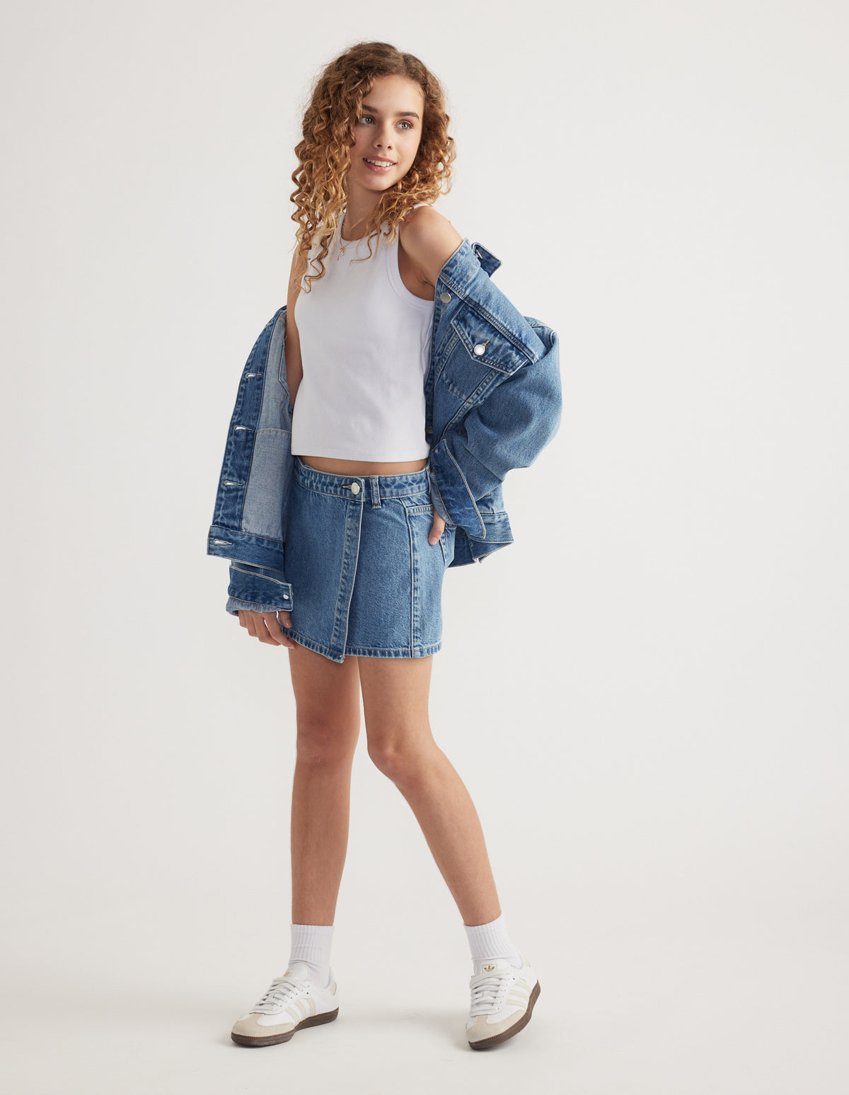Girl wearing a blue denim skort with a white crop top, oversized denim jacket and sneakers