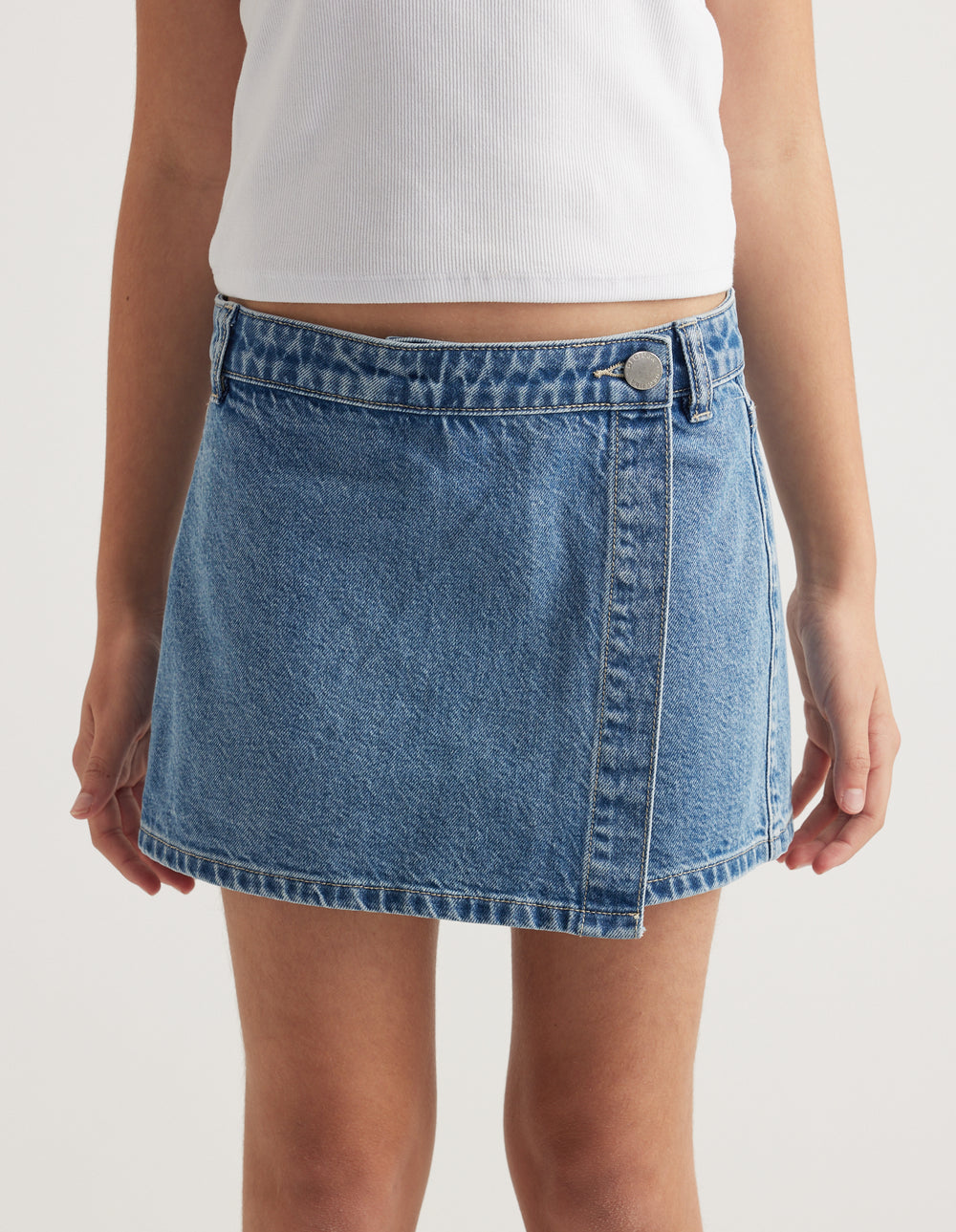Girl wearing a light blue denim skort with a wrap front and button closure, paired with a white tank top