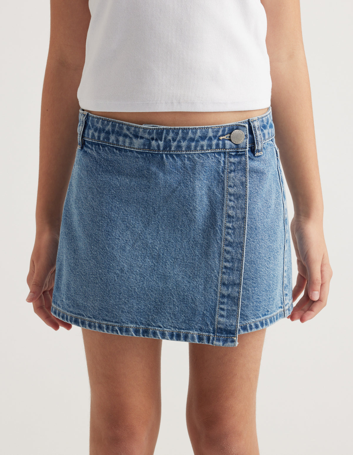 Girl wearing a light blue denim skort with a wrap front and button closure, paired with a white tank top