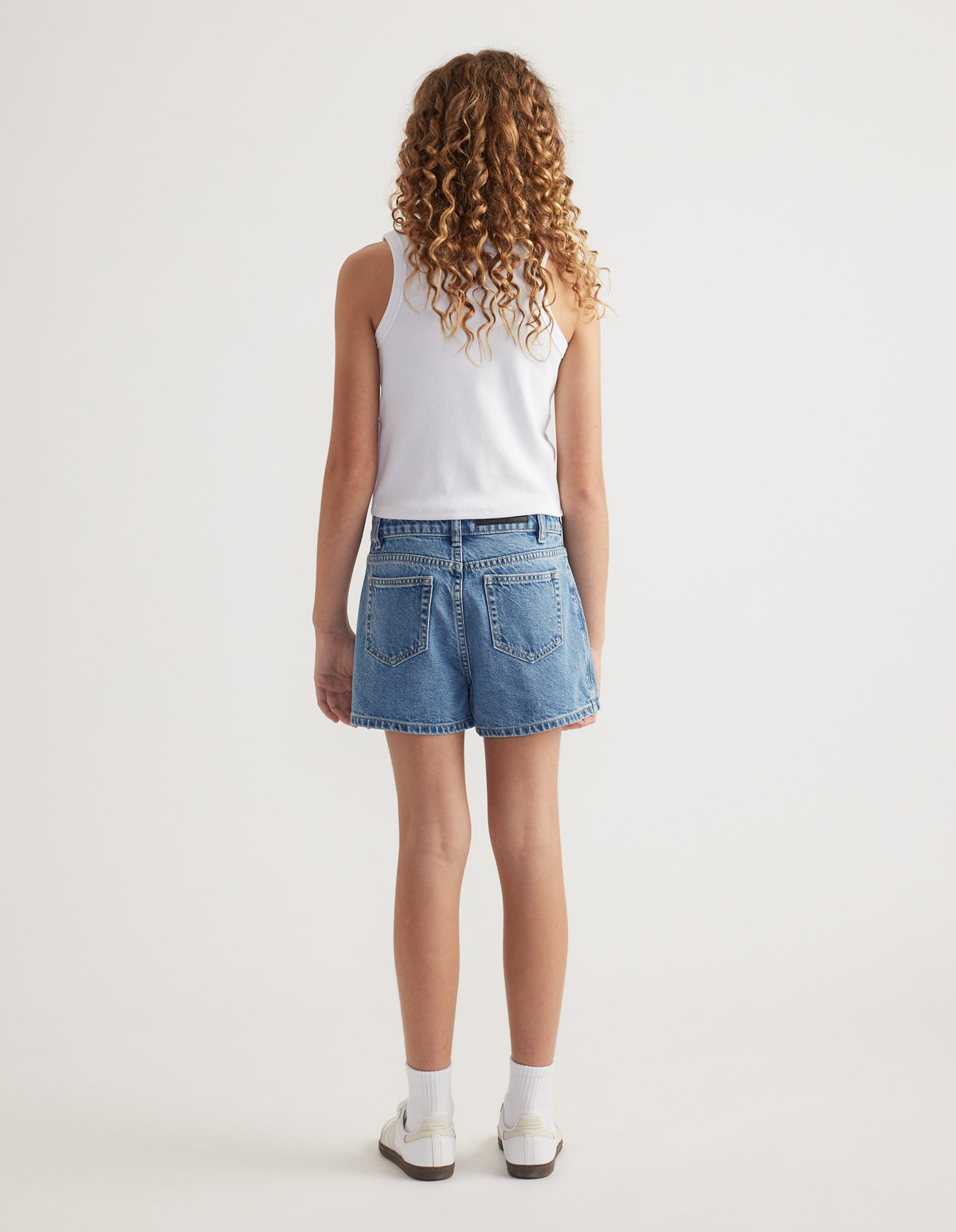 Back view of girl wearing a blue denim skort with a white tank top, ankle socks and sneakers