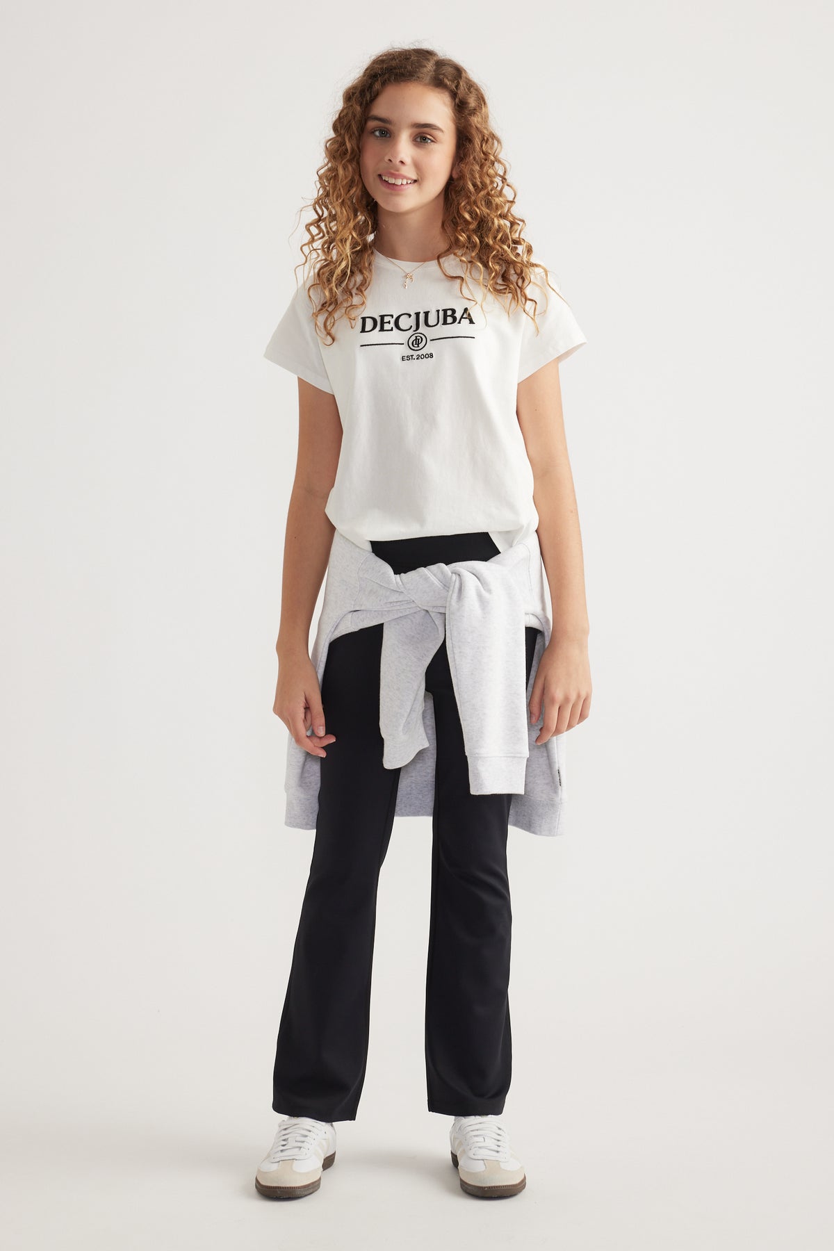 Girl wearing a white heritage graphic tee with black pants, a sweatshirt tied at the waist and sneakers