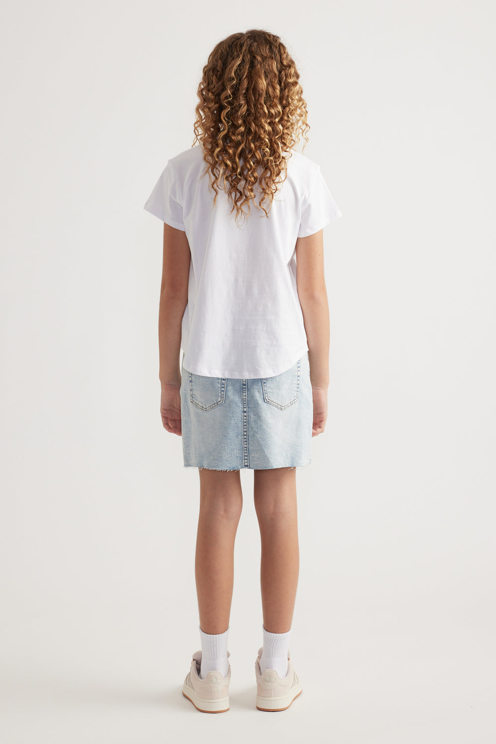 Back view of girl wearing a white heritage graphic tee with a curved hem, denim skirt and sneakers