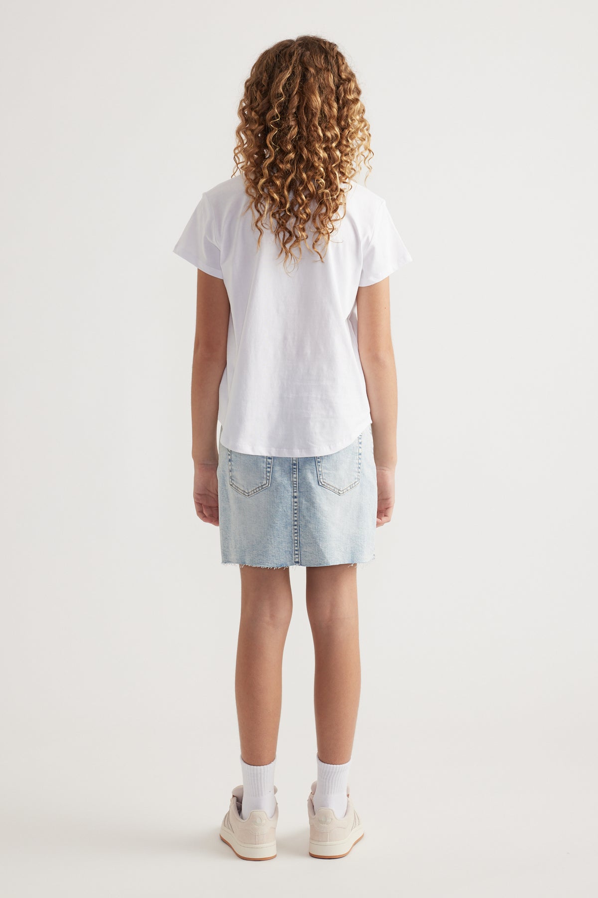 Back view of girl wearing a white heritage graphic tee with a curved hem, denim skirt and sneakers