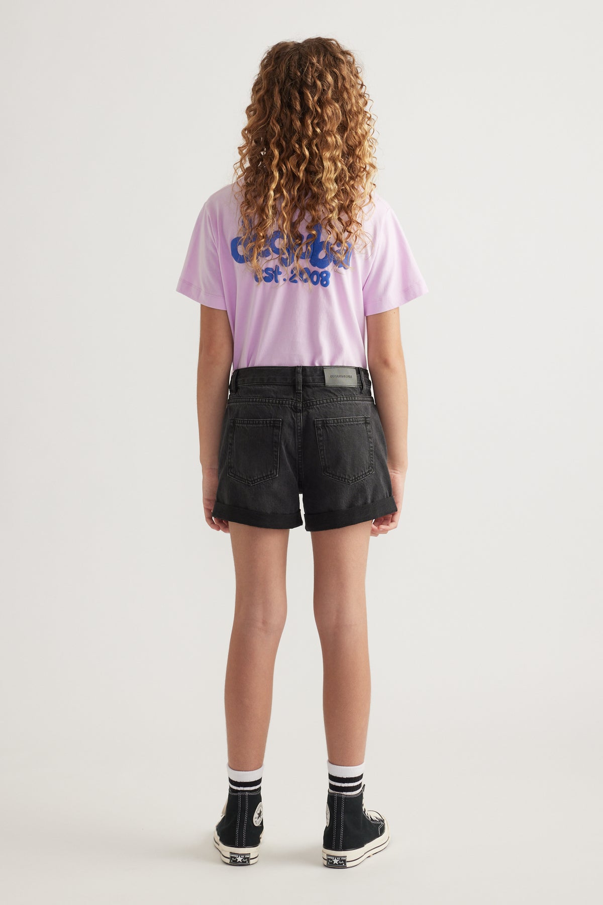 Back view of a girl wearing black denim shorts with a pink graphic tee and high top sneakers
