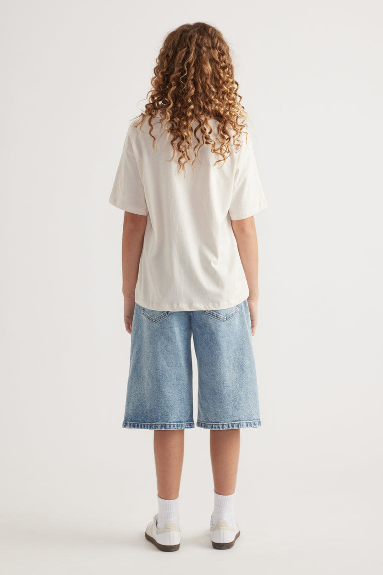 Back view of girl in an oversized cream tee, light wash knee-length denim shorts and sneakers
