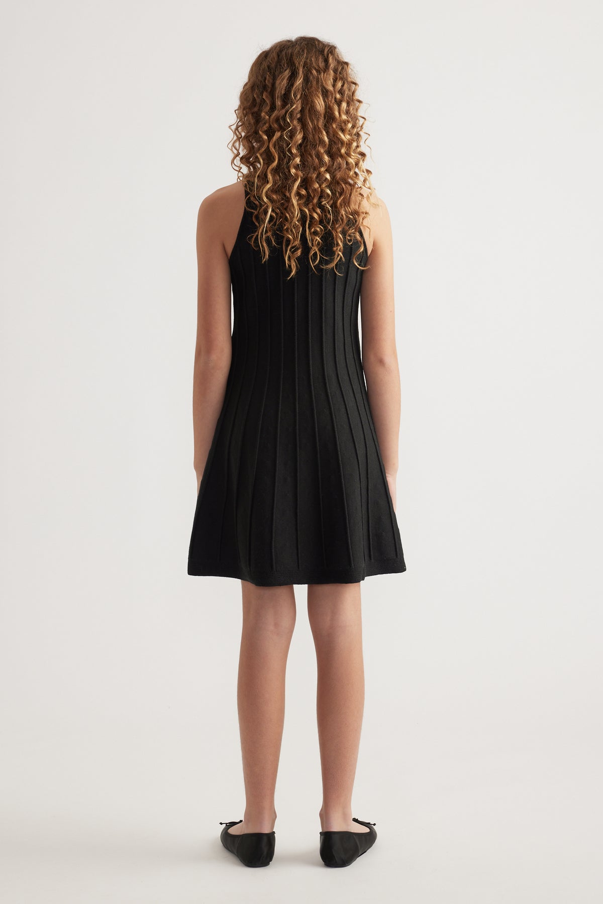 Girl seen from behind wearing a black sleeveless knit fit and flare mini dress with black flats