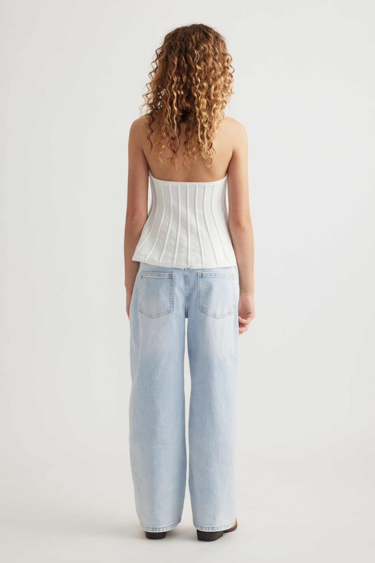 Back view of a girl wearing a white halter top with light blue wide leg jeans and black shoes