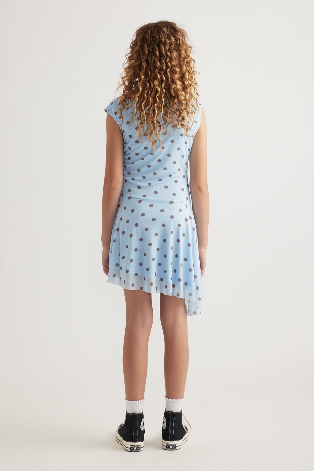 Girl viewed from behind wearing a blue polka dot asymmetric dress with black sneakers